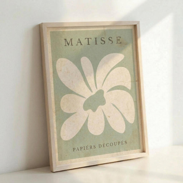 May include: Framed art print in light wood frame leaning against white wall. Green background with large white flower cutout design in Matisse's paper collage style. Text reads "MATISSE" at top and "PAPIERS DÉCOUPÉS" at bottom in dark letters. Simple daisy-like bloom with rounded petals and organic center shape fills most of the composition.