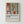 May include: Framed poster in light wood frame hanging on white wall, featuring Matisse-inspired artwork with abstract interior scene showing potted plant with green leaves and orange flowers, terracotta vases on shelves, geometric shapes in warm earth tones of rust, pink, and blue. Bold black text below reads "Celebrating Colors of Life" with smaller descriptive text underneath.