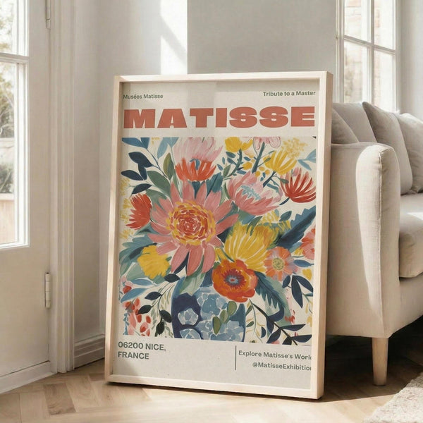 May include: A framed poster featuring a vibrant floral bouquet in Matisse-inspired style with coral, yellow, and blue blooms against a cream background. The artwork displays "MATISSE" in bold red letters at the top and "06200 NICE, FRANCE" at the bottom. The light wood frame leans against a white wall next to a beige upholstered chair in a bright, naturally-lit room with white window frames.