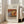 May include: Framed art print leaning against white wall beside beige upholstered chair in bright living room with large windows. The poster features a Renoir still life painting of oranges, bananas, and blue-patterned teacup on warm wooden surface. Cream-colored mat with black text above and below the artwork. Natural wood frame, approximately 2-3 feet tall, casting soft shadows on light wooden floor.