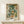 May include: Framed botanical art print featuring William Morris-inspired floral design with cream peonies, sage green foliage, and orange accent flowers against dark background, displayed on light wood console table beside small wrapped gift with twine and sprig decoration against neutral wall