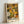 Framed art print featuring abstract floral pattern in warm earth tones including yellows, oranges, greens, and browns. The design shows overlapping stylized flowers and dotted circular patterns creating a dense, psychedelic composition. Text at bottom reads "YAYOI KUSAMA" in black letters and "NAGOYA" in yellow, with Japanese characters below. Print is displayed in light wooden frame leaning against white wall.