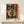 May include: Framed cubist-style artwork displaying abstract faces with geometric shapes in vibrant blues, oranges, yellows and reds, leaning against white wall on light wooden console table. Small wrapped gift with twine sits beside the poster. The artwork features multiple overlapping facial profiles with distinctive eyes and lips in Picasso-inspired cubist style.