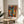 May include: Colorful cubist-style artwork featuring abstract faces with geometric shapes in blues, oranges, reds, and yellows, displayed in thin wooden frame on round wooden side table next to beige upholstered chair. Sunlight streams across white wall creating diagonal shadows, with green plant leaves visible in corner of modern living room setting.