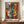 May include: Framed Picasso-style cubist artwork featuring abstract faces in vibrant blues, oranges, yellows, and reds, displayed on wooden sideboard next to white ceramic vase and stacked books in elegant room with white paneled walls