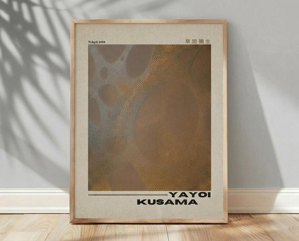May include: Framed poster with light wood frame leaning against white wall on wooden floor. Palm leaf shadows cast across wall. Poster features brown and beige abstract dotted pattern with "Tōkyō 1998" and Japanese characters at top, "YAYOI KUSAMA" at bottom in black text. Cream colored mat border surrounds artwork. Natural lighting creates geometric shadow patterns in minimalist room setting.