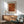 May include: Framed orange and brown dotted abstract art print hanging on white wall above wooden sideboard with table lamp. Mid-century modern living room setting with sage green upholstered armchair, round coffee table, and natural sunlight casting shadows. Print features textured dot pattern in warm rust tones with Japanese characters and artist attribution text below.