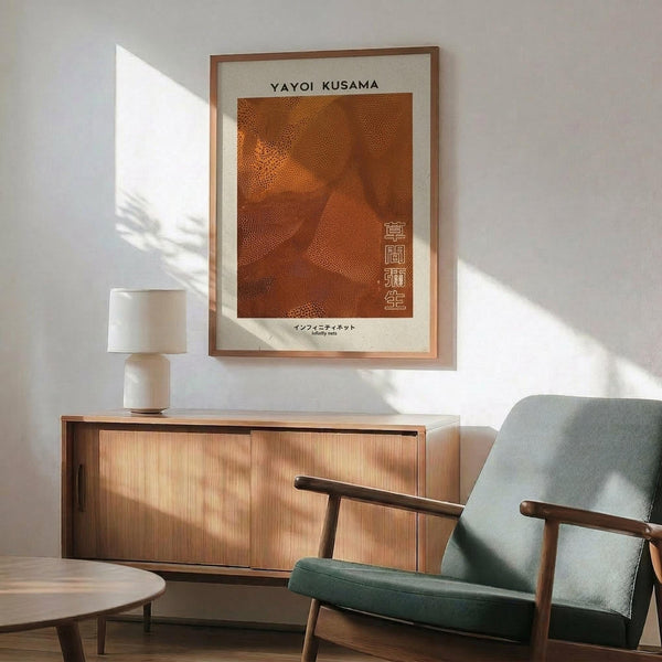 May include: Framed orange and brown dotted abstract art print hanging on white wall above wooden sideboard with table lamp. Mid-century modern living room setting with sage green upholstered armchair, round coffee table, and natural sunlight casting shadows. Print features textured dot pattern in warm rust tones with Japanese characters and artist attribution text below.
