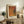 May include: Abstract art poster with warm orange, burgundy, and teal dotted pattern in light wood frame, displayed on rustic wooden sideboard beside fern plant, books, and ceramic vase in softly lit living room with beige armchair and neutral walls