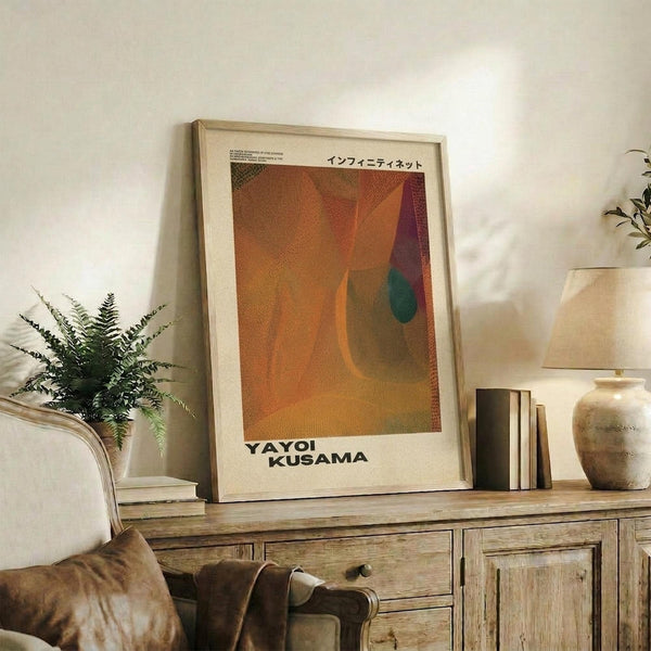May include: Abstract art poster with warm orange, burgundy, and teal dotted pattern in light wood frame, displayed on rustic wooden sideboard beside fern plant, books, and ceramic vase in softly lit living room with beige armchair and neutral walls