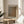 May include: Framed art print with abstract brown and beige dotted pattern displayed on round wooden side table next to beige upholstered chair. Natural sunlight streams through window creating soft shadows on white wall. Green plant leaves visible in corner of minimalist living room setting.