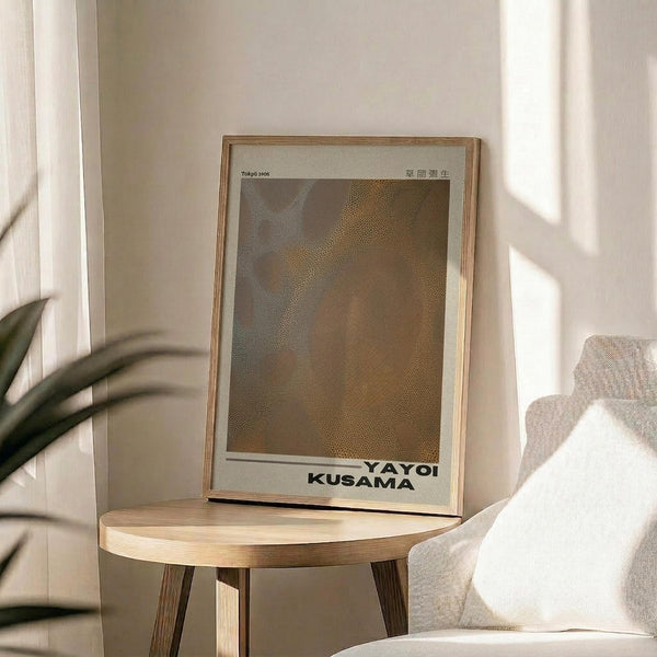 May include: Framed art print with abstract brown and beige dotted pattern displayed on round wooden side table next to beige upholstered chair. Natural sunlight streams through window creating soft shadows on white wall. Green plant leaves visible in corner of minimalist living room setting.