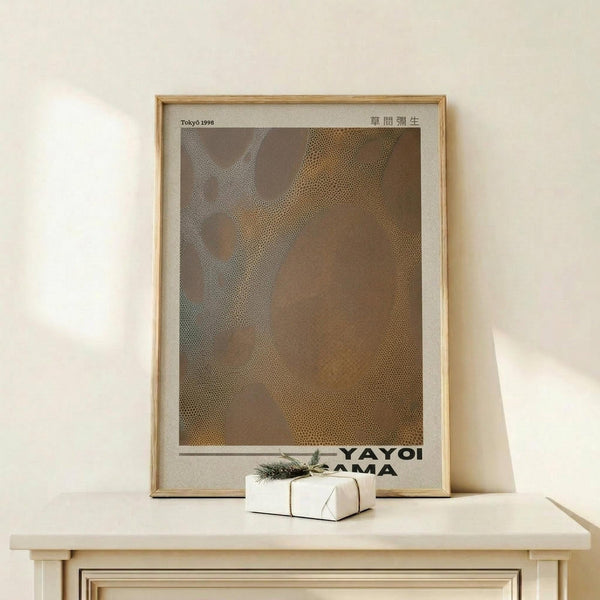 May include: Framed art print leaning against cream wall on light wood console table, featuring abstract brown and beige dotted pattern with "Tokyo 1998" and Japanese characters at top, "YAYOI KUSAMA" text at bottom. Small white gift box with greenery sits in front. Natural wood frame with white matting.