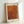 May include: Framed art print in light wooden frame leaning against white wall. Orange and rust colored abstract pattern with flowing dots and curves in Kusama's signature infinity nets style. Japanese text characters visible on right side of artwork. Print has cream colored border with black text at top and bottom in both English and Japanese characters.