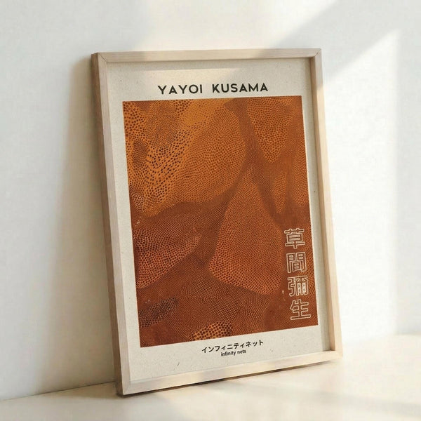 May include: Framed art print in light wooden frame leaning against white wall. Orange and rust colored abstract pattern with flowing dots and curves in Kusama's signature infinity nets style. Japanese text characters visible on right side of artwork. Print has cream colored border with black text at top and bottom in both English and Japanese characters.