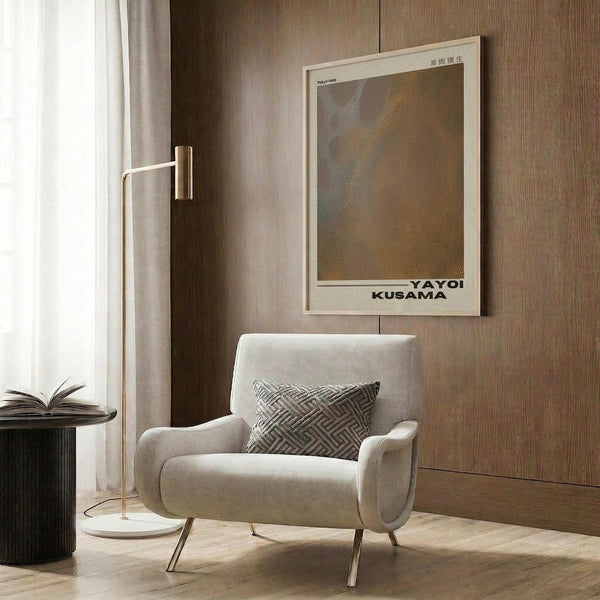 May include: Framed Yayoi Kusama Tokyo 1998 poster with abstract dotted pattern in golden and gray tones, displayed on wood-paneled wall above cream upholstered armchair with geometric patterned pillow. Modern brass floor lamp and dark round side table with open book complete the neutral-toned reading corner setting.