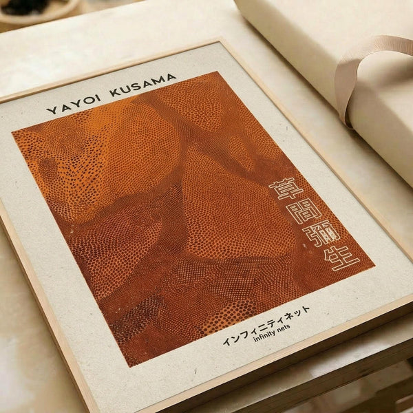 Orange halftone dot pattern artwork featuring abstract organic forms reminiscent of infinity nets, displayed on cream paper with Japanese text vertically arranged on right side. The piece shows varying densities of dots creating flowing, cellular-like shapes in warm rust and orange tones. Mounted print lies flat on neutral surface alongside rolled paper tubes.