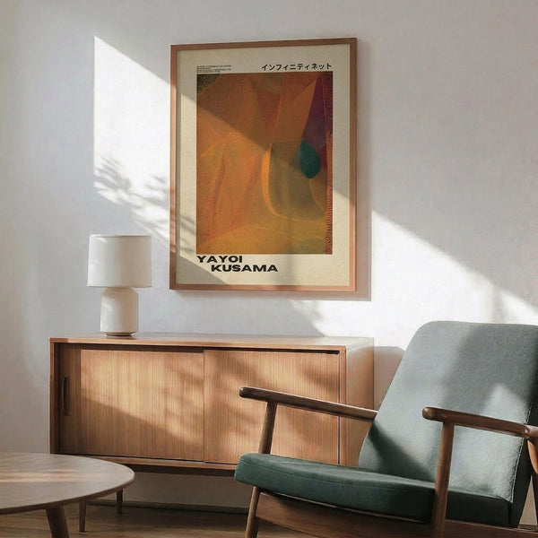 May include: Framed art poster with orange, yellow, and purple abstract dotted design hanging on white wall above wooden sideboard. Mid-century modern living room setting with sage green upholstered chair, white ceramic table lamp, and round coffee table. Warm natural lighting creates geometric shadows across the scene.