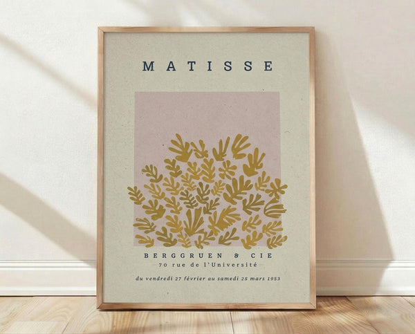 May include: Framed art print featuring Matisse exhibition poster with golden botanical cutout design on pink and cream background, displayed against white textured wall on light wood floor with natural lighting creating soft shadows