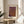 May include: Framed art print with abstract brown and purple textured pattern resembling organic networks or cellular structures, displayed on round wooden side table next to beige upholstered chair. Natural lighting creates soft shadows across cream colored wall. Green plant leaves visible in corner of minimalist interior setting.