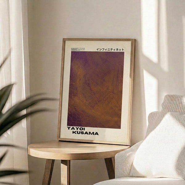 May include: Framed art print with abstract brown and purple textured pattern resembling organic networks or cellular structures, displayed on round wooden side table next to beige upholstered chair. Natural lighting creates soft shadows across cream colored wall. Green plant leaves visible in corner of minimalist interior setting.