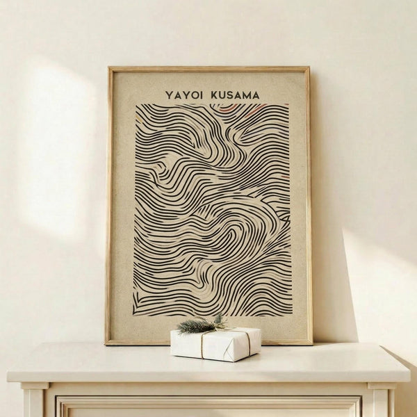 May include: Framed poster with flowing black wavy lines creating organic ripple patterns on cream background, displayed on light wood furniture beside small white wrapped gift with green sprig. Artwork features rhythmic curved stripes that undulate across the surface in a hypnotic wave-like design. Natural wood frame complements the minimalist neutral color palette in this clean, bright interior setting.