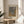 May include: Framed poster with dotted abstract pattern in gray and golden yellow tones, displayed on light wood round side table next to beige upholstered chair. Japanese text visible at top of artwork. Soft natural lighting creates shadows on neutral wall background, with plant leaves partially visible in corner of minimalist room setting.