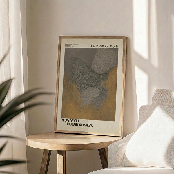 May include: Framed poster with dotted abstract pattern in gray and golden yellow tones, displayed on light wood round side table next to beige upholstered chair. Japanese text visible at top of artwork. Soft natural lighting creates shadows on neutral wall background, with plant leaves partially visible in corner of minimalist room setting.