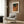 May include: Abstract art poster with name "Yayoi Kusama" at top, featuring flowing organic shapes in teal, orange, brown and gold tones with dotted textures, displayed in light wood frame on wood-paneled wall above cream upholstered armchair with geometric patterned pillow, beside brass floor lamp and dark side table with open book