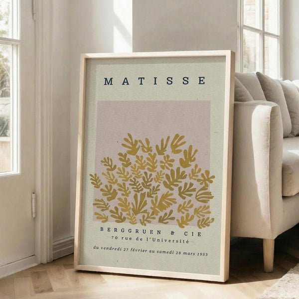 May include: Framed vintage-style art print leaning against white wall in bright living room with beige sofa. Poster features golden yellow botanical leaf pattern on pink background with "MATISSE" text at top and French gallery text below. Light wood frame, natural lighting from windows.