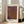 May include: Framed art print leaning against white wall next to beige sofa in bright living room. The poster features a textured gradient pattern in purple, brown, and gold tones with dotted and web-like designs. Japanese text appears at top, with "YAYOI KUSAMA" in bold black letters at bottom. Natural wood frame, approximately 2-3 feet tall, positioned on light wooden floor near large windows.