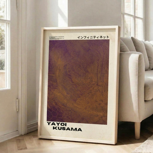 May include: Framed art print leaning against white wall next to beige sofa in bright living room. The poster features a textured gradient pattern in purple, brown, and gold tones with dotted and web-like designs. Japanese text appears at top, with "YAYOI KUSAMA" in bold black letters at bottom. Natural wood frame, approximately 2-3 feet tall, positioned on light wooden floor near large windows.