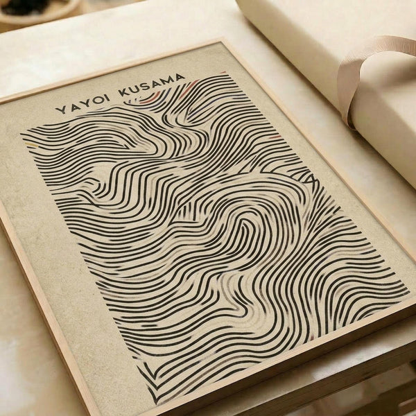A minimalist art print featuring flowing, wavy black lines creating an abstract topographical pattern on cream-colored paper. The organic curves and contours resemble fingerprint ridges or topographic map lines, creating a mesmerizing optical effect. The artwork is displayed in a simple light wood frame, photographed at an angle on a neutral beige surface with soft natural lighting and cream-colored ribbons visible in the composition.