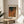 May include: Framed art print on wooden side table next to beige upholstered chair and plant. Print features abstract design with overlapping curved shapes in warm earth tones - oranges, browns, greens - with dotted texture patterns. Text at top reads "Yayoi Kusama" with Japanese characters below. Natural wood frame matches round table. Soft natural lighting creates shadows on neutral wall background.
