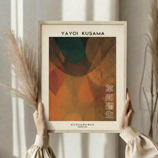May include: Person holding framed art print featuring abstract overlapping circular forms in warm orange, brown, and green tones with dotted texture patterns. Light wood frame with cream matting displays Japanese text. Dried pampas grass visible to left, neutral beige clothing and soft window lighting create minimalist aesthetic.