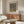 May include: Framed Yayoi Kusama exhibition poster with abstract dotted patterns in grey and golden yellow tones, displayed above a mid-century modern walnut credenza with brass hardware. White sculptural chair, glass-top coffee table, and brass floor lamp create a minimalist Scandinavian interior with cream walls and natural lighting.