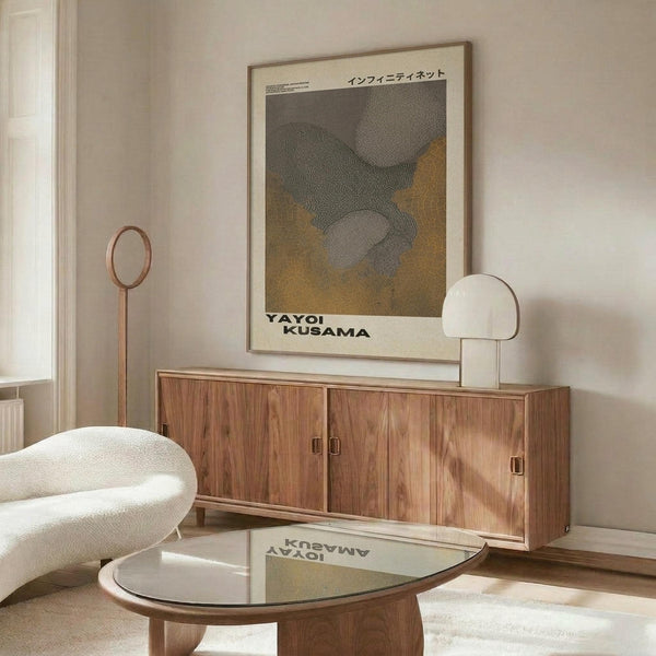 May include: Framed Yayoi Kusama exhibition poster with abstract dotted patterns in grey and golden yellow tones, displayed above a mid-century modern walnut credenza with brass hardware. White sculptural chair, glass-top coffee table, and brass floor lamp create a minimalist Scandinavian interior with cream walls and natural lighting.