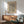 May include: Framed art print with flowing black wavy lines on cream background, displayed above wooden sideboard with white ceramic lamp. Mid-century modern living room setting features sage green upholstered chair with wooden arms, round coffee table, and natural sunlight casting shadows on white wall.