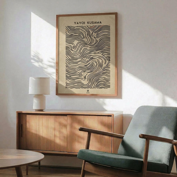May include: Framed art print with flowing black wavy lines on cream background, displayed above wooden sideboard with white ceramic lamp. Mid-century modern living room setting features sage green upholstered chair with wooden arms, round coffee table, and natural sunlight casting shadows on white wall.