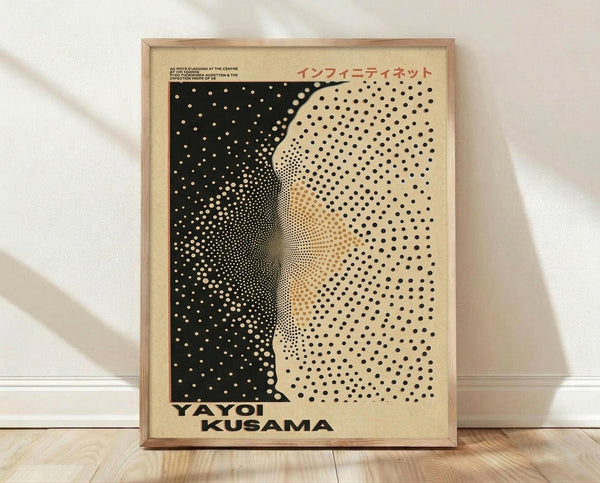 May include: Framed art print leaning against white wall on light wooden floor. Black and cream poster features abstract dotted pattern creating curved, wave-like forms reminiscent of polka dot infinity nets. Japanese text appears in red at top, artist name in bold black letters at bottom. Natural wood frame with warm lighting casting soft shadows on textured white wall.