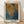 May include: Framed poster print leaning against white wall on wooden floor. Light wooden frame contains artwork with organic dotted patterns in golden yellow, brown, and deep blue tones creating flowing, net-like textures. Text reads "Tokyo 1998" at top with Japanese characters, "YAYOI KUSAMA" at bottom. Natural lighting casts soft shadows on cream-colored wall behind the minimalist display.
