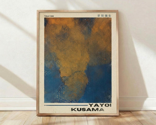 May include: Framed poster print leaning against white wall on wooden floor. Light wooden frame contains artwork with organic dotted patterns in golden yellow, brown, and deep blue tones creating flowing, net-like textures. Text reads "Tokyo 1998" at top with Japanese characters, "YAYOI KUSAMA" at bottom. Natural lighting casts soft shadows on cream-colored wall behind the minimalist display.