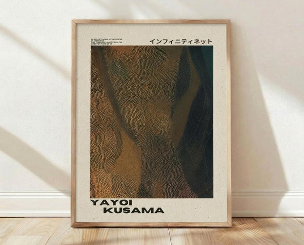 May include: A framed art print leaning against a white wall on light wood flooring. The print features an abstract dotted pattern in earthy tones of brown, green, and gold with flowing, organic forms. Japanese text appears at the top, with "YAYOI KUSAMA" in bold black letters at the bottom. The piece has a cream-colored background and is displayed in a thin natural wood frame. Soft natural lighting creates gentle shadows on the textured white wall.