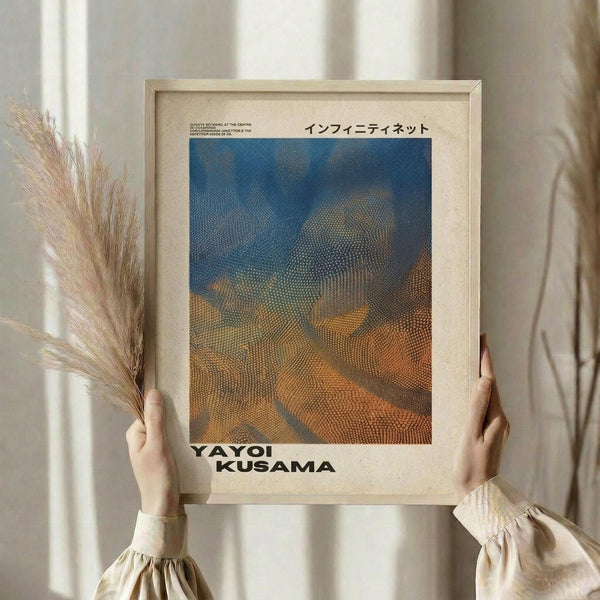 May include: Hands holding a framed art print with Japanese text and a dotted pattern artwork in blue and orange tones, alongside dried pampas grass stems. The wooden frame contains a halftone-style image resembling organic, flowing forms. Person wears a cream-colored top with gathered sleeves against soft natural lighting and neutral curtains in the background.