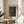 May include: Framed art print displaying abstract dotted pattern in earthy tones of brown, green, and black with Japanese text at top and artist name at bottom. Print sits on round wooden side table next to beige upholstered chair with decorative pillow. Soft natural lighting creates shadows on neutral wall, with plant leaves visible in corner of minimalist interior setting.