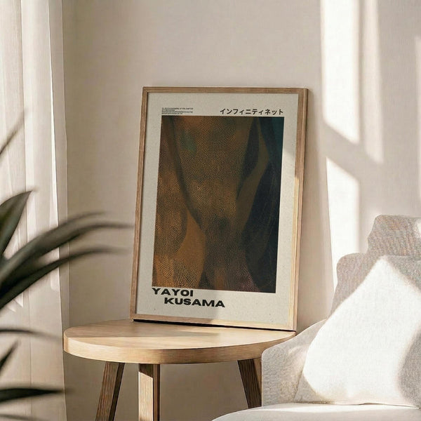 May include: Framed art print displaying abstract dotted pattern in earthy tones of brown, green, and black with Japanese text at top and artist name at bottom. Print sits on round wooden side table next to beige upholstered chair with decorative pillow. Soft natural lighting creates shadows on neutral wall, with plant leaves visible in corner of minimalist interior setting.