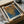 May include: Abstract art print featuring orange and blue dotted patterns reminiscent of cellular or net-like structures, displayed in black frame with white matting. Print sits in open cardboard shipping box surrounded by white tissue paper and wooden dowels on neutral surface. Artwork shows organic flowing forms created with small dots in warm golden orange transitioning to deep blue tones.