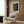 May include: Modern reading nook with beige upholstered armchair featuring geometric patterned pillow, gold floor lamp with white circular base, dark round side table with open book, and framed art poster on wood-paneled wall. The poster displays abstract dotted patterns in earthy tones of brown, green, and orange with Japanese text and artist name at bottom in light wooden frame.