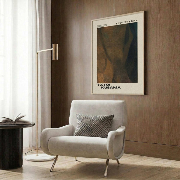 May include: Modern reading nook with beige upholstered armchair featuring geometric patterned pillow, gold floor lamp with white circular base, dark round side table with open book, and framed art poster on wood-paneled wall. The poster displays abstract dotted patterns in earthy tones of brown, green, and orange with Japanese text and artist name at bottom in light wooden frame.