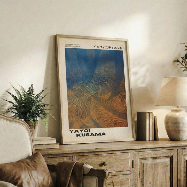 May include: A framed art print displaying an abstract dotted pattern in blue, orange and earth tones, styled in a living room setting on a wooden sideboard alongside books, a ceramic vase, and fern plant. The poster features Japanese text at the top and "YAYOI KUSAMA" text at the bottom, presented in a light wood frame against white walls with warm natural lighting.